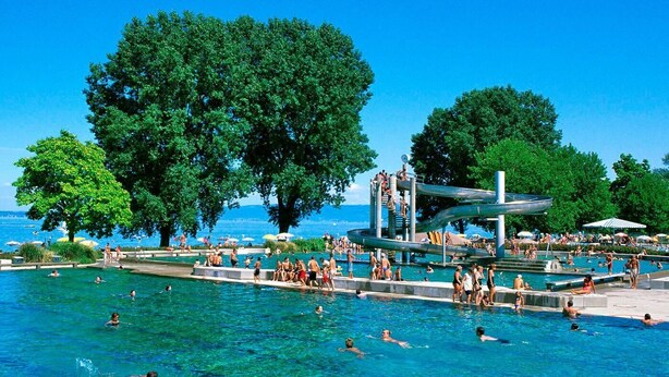 Una piscina pubblica con diverse corsie, circondata da alberi e un cielo azzurro terso.