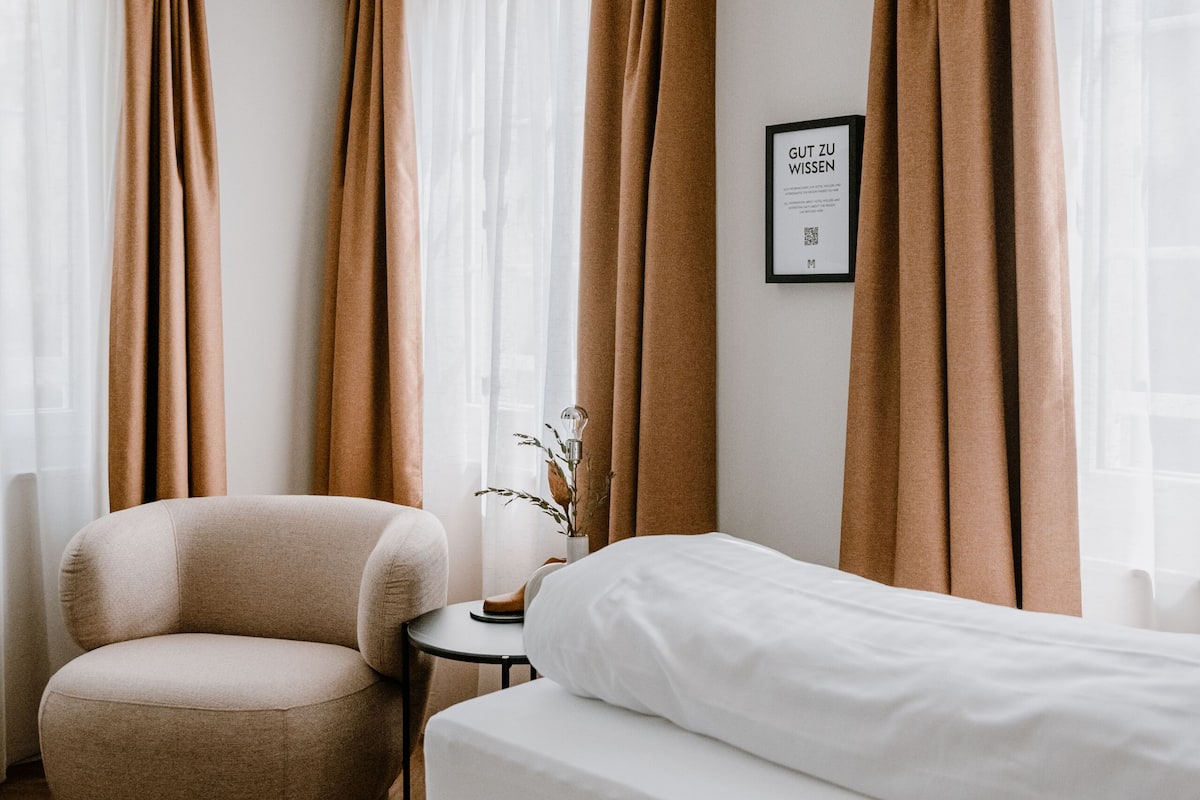 A hotel room with a bed, a round armchair, a small table with a vase, and a framed sign on the wall.