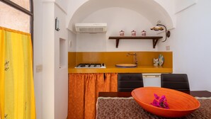 A compact kitchen with a gas stove, a sink, and a small countertop. There is a dining table with a bowl of flowers and two chairs. - Appartamento Dammuso Anita - Iso... (Isola di pantelleria)