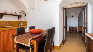 A dining area with a wooden table, chairs, and a central orange bowl. There is a small shelf with decorative items above the table. The room has white walls and a wooden door leading to another room. - Appartamento Dammuso Anita - Iso... (Isola di pantelleria)