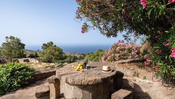 Une table en pierre, sur laquelle se trouvent une carafe, des verres et un chapeau, le tout entouré de verdure et offrant une vue sur la mer.