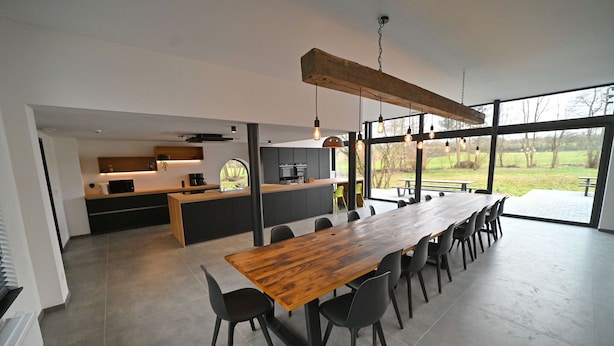 Una cocina moderna con una isla grande, un comedor con una larga mesa de madera y sillas, y ventanas amplias que ofrecen vistas al exterior.