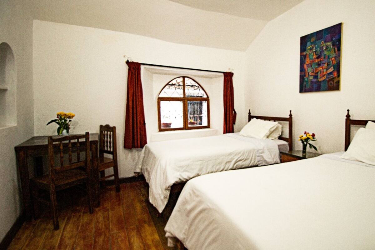 A room with two beds, a wooden desk, a chair, a window with curtains, and a painting on the wall.
