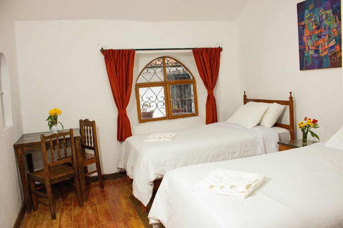 A bedroom with two beds, a window with curtains, a wooden chair, and a table with a vase of flowers.
