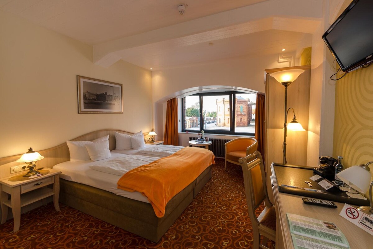 Habitación de hotel con cama, mesitas de noche, escritorio, silla, televisor y ventana con vistas a edificios.