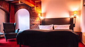 Habitación de hotel con pared de ladrillo, una cama grande con ropa blanca, un sillón oscuro y dos lámparas fijadas a la pared.