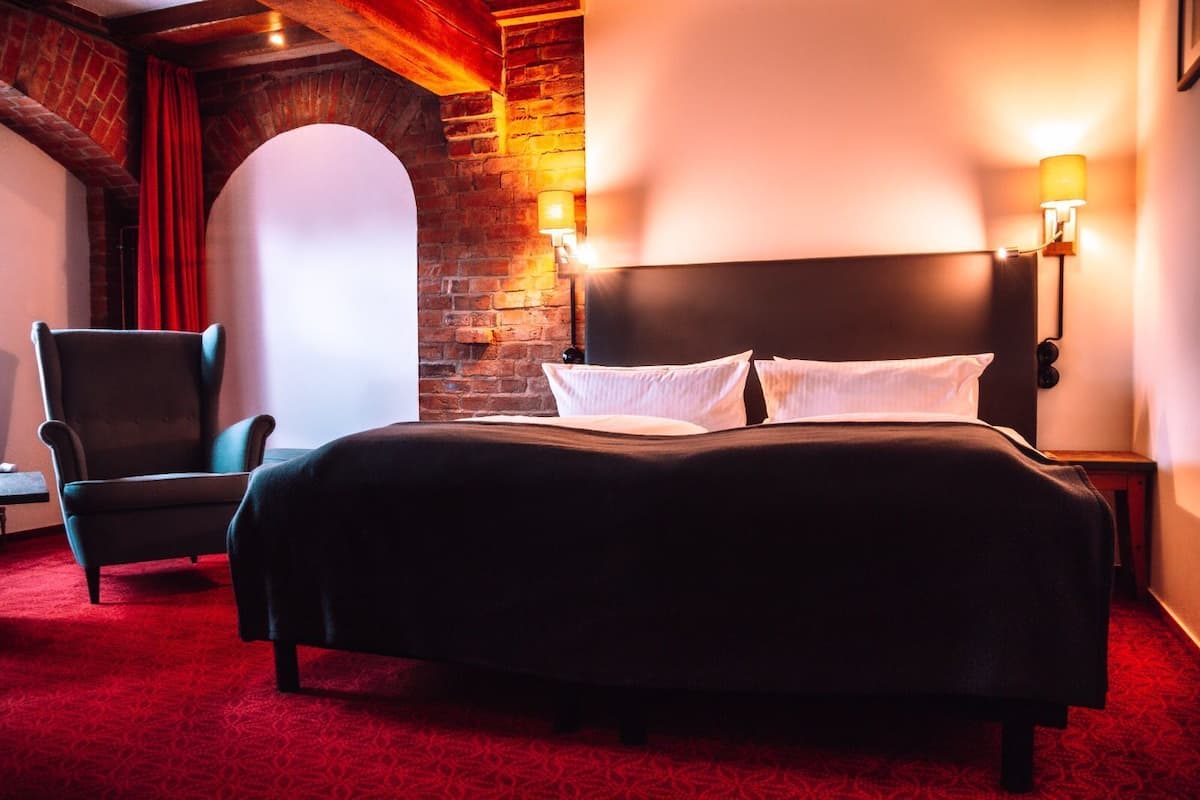 A hotel room with a brick wall, a large bed with white linens, a dark armchair, and two wall-mounted lamps.