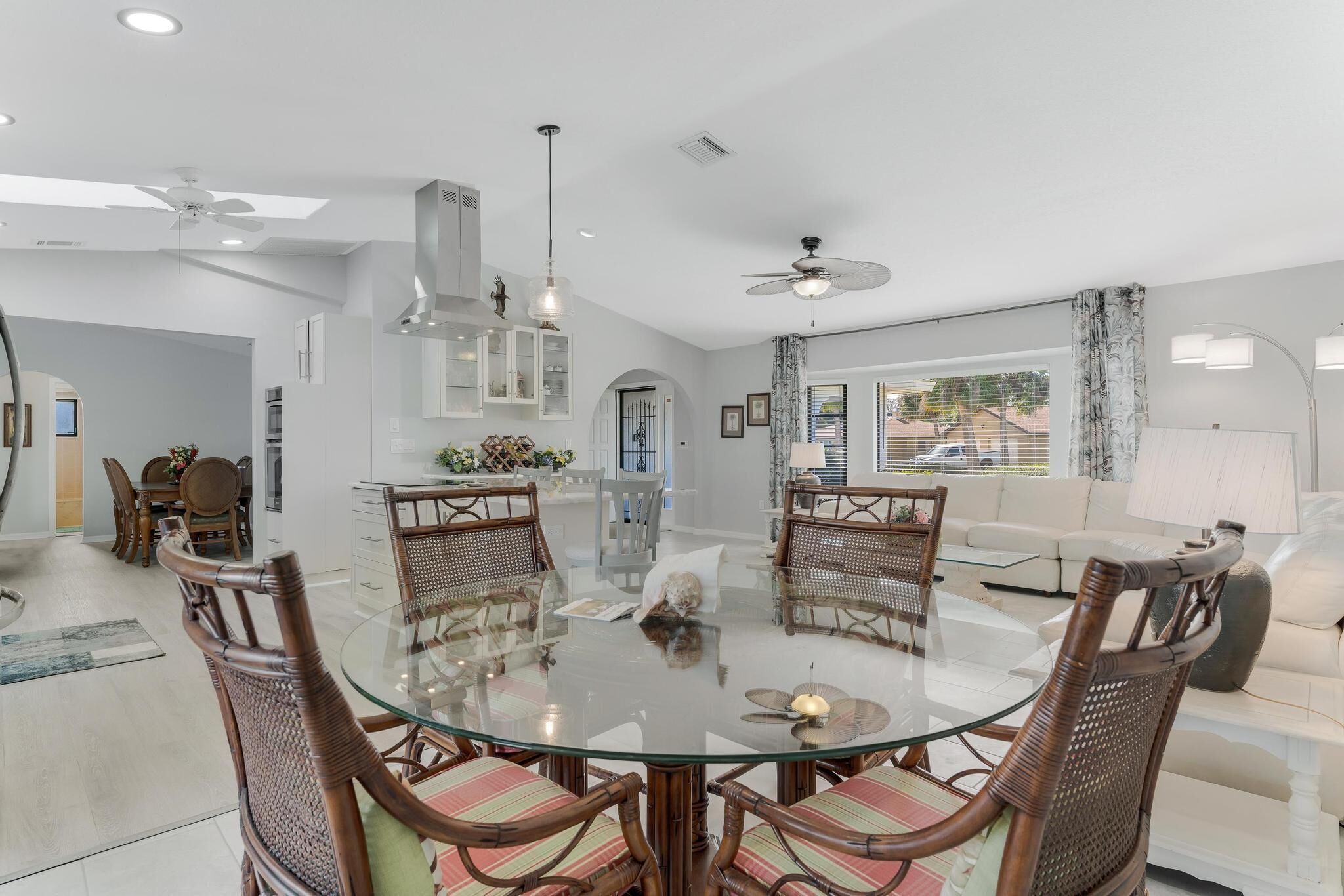A spacious dining area with a round glass table surrounded by wicker chairs, a modern kitchen in the background, and a large window offering a view of the outside.