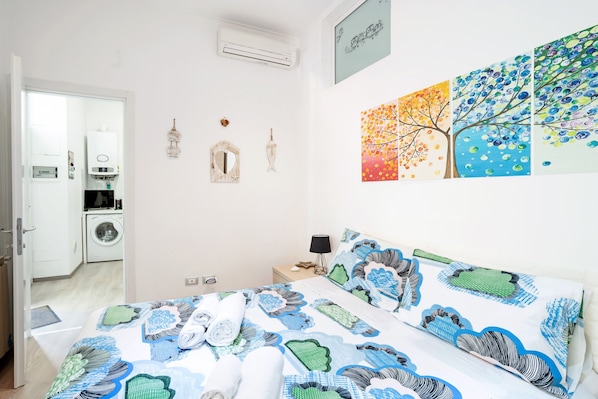 Une chambre à coucher comprenant un lit, une table de chevet, un climatiseur fixé au mur, un lave-linge et une porte donnant sur une autre pièce.