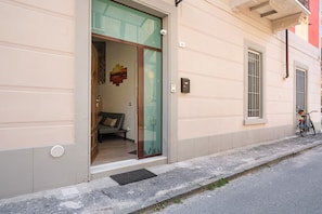 Une entrée d’immeuble avec une porte en verre, un intérieur visible avec un canapé, et un vélo garé à l’extérieur.