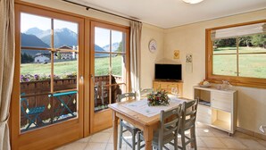 A room with a wooden table, chairs, a TV, and a view of mountains through large windows.