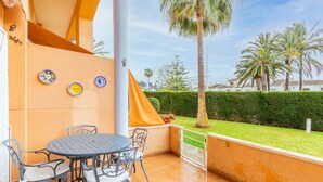 A patio with a round table and chairs, a yellow awning, and a view of a well-maintained garden with palm trees. - Pet-Friendly Apartment 'Edificio Menorca' close to the Sea with Shared Pool & Wi-Fi (Alicante)