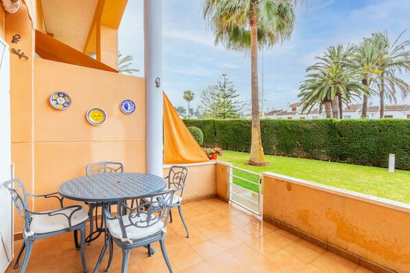 A patio with a round table and chairs, a yellow awning, and a view of a well-maintained garden with palm trees.