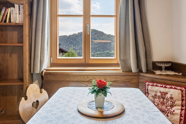 A room with a wooden bookshelf, a table with a white tablecloth, a vase with a red flower, and a window with a view of a hill.