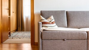 A grey sofa with a patterned throw pillow, a wooden floor, and a wooden door in the background. - Holiday Apartment 'Haus Thurn Apartment B' with Mountain View, Garden & Wi-Fi (Kastelruth)