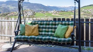 Un fauteuil suspendu doté d’un coussin et de plusieurs oreillers à carreaux, installé sur une terrasse en pierre offrant une vue sur un village et des montagnes.