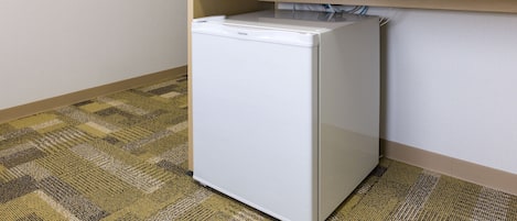 A white refrigerator in a corner of a room with a patterned carpet and a wooden shelf above it.