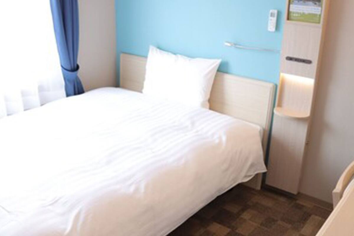 Een net opgemaakt bed in een hotelkamer met een geruit vloertje, een blauwe wand en een houten kastje met verlichting.