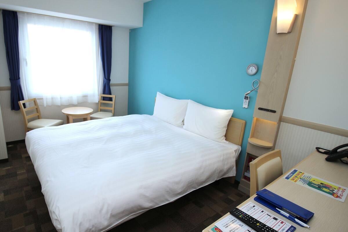 Een hotelkamer met een bed, een bureau met telefoon en afstandsbediening, een stoel, een klein tafeltje en een raam met gordijnen.