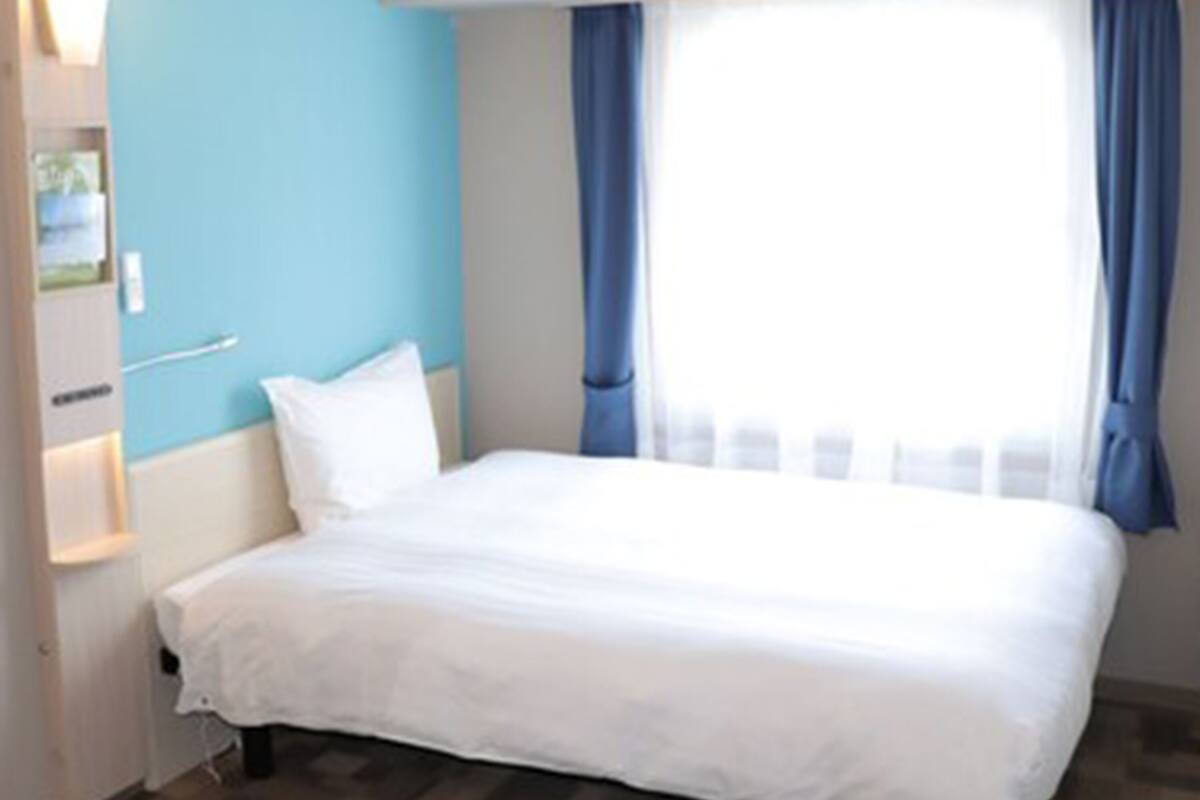 Hotelkamer met een bed, nachtkastje, raam met blauwe gordijnen en een wand met lichtschakelaar.
