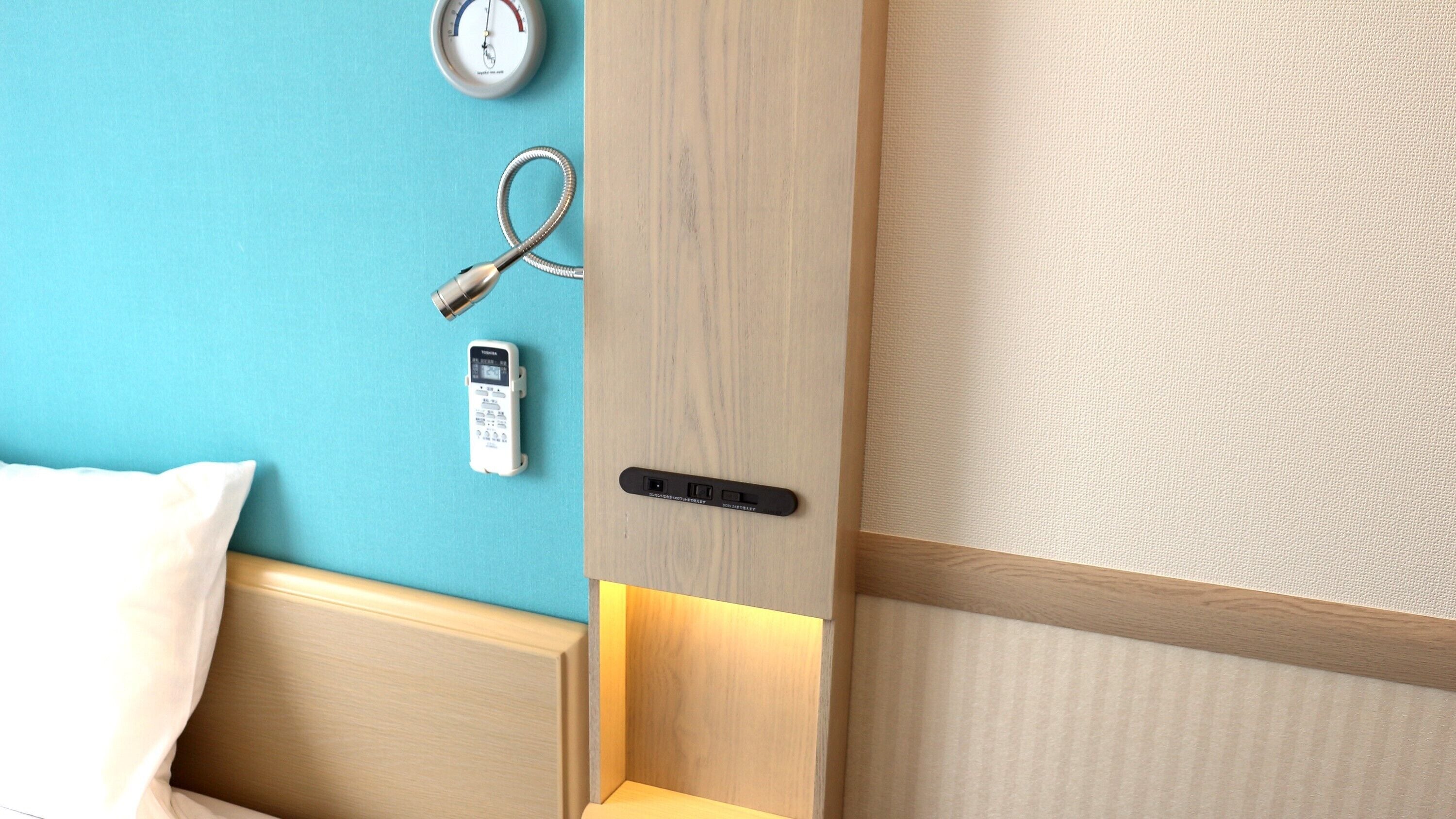 A hotel room with a wooden bed frame, a bedside table with a lamp, a wall-mounted alarm clock, and a remote control.