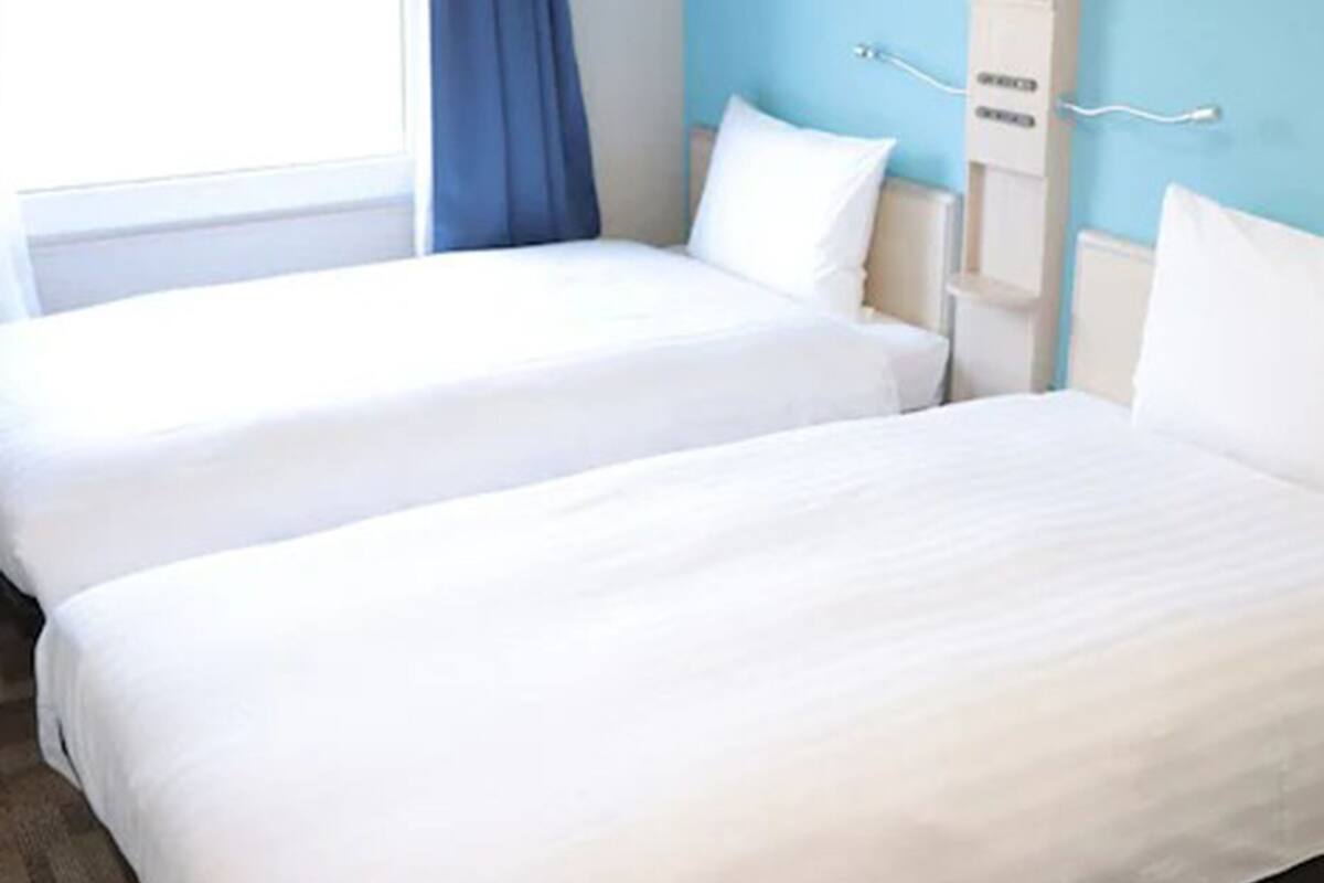 Een hotelkamer met twee eenpersoonsbedden, een raam met een blauw gordijn en een witte muur met een thermostaat.