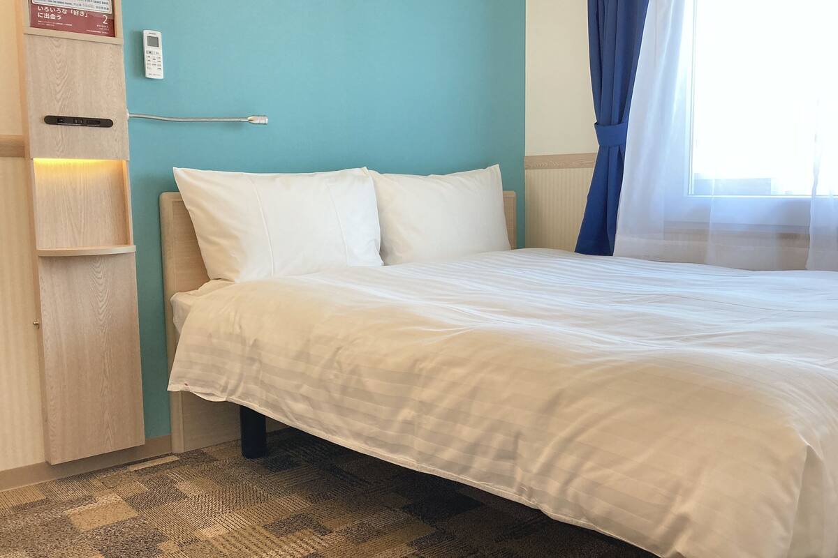 Een net opgemaakt bed met witte lakens in een kamer met teal muren, een zichtbare thermostaat en een raam met dunne gordijnen.