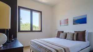 A bedroom with a bed, a window, a desk with a lamp, and two paintings on the wall.