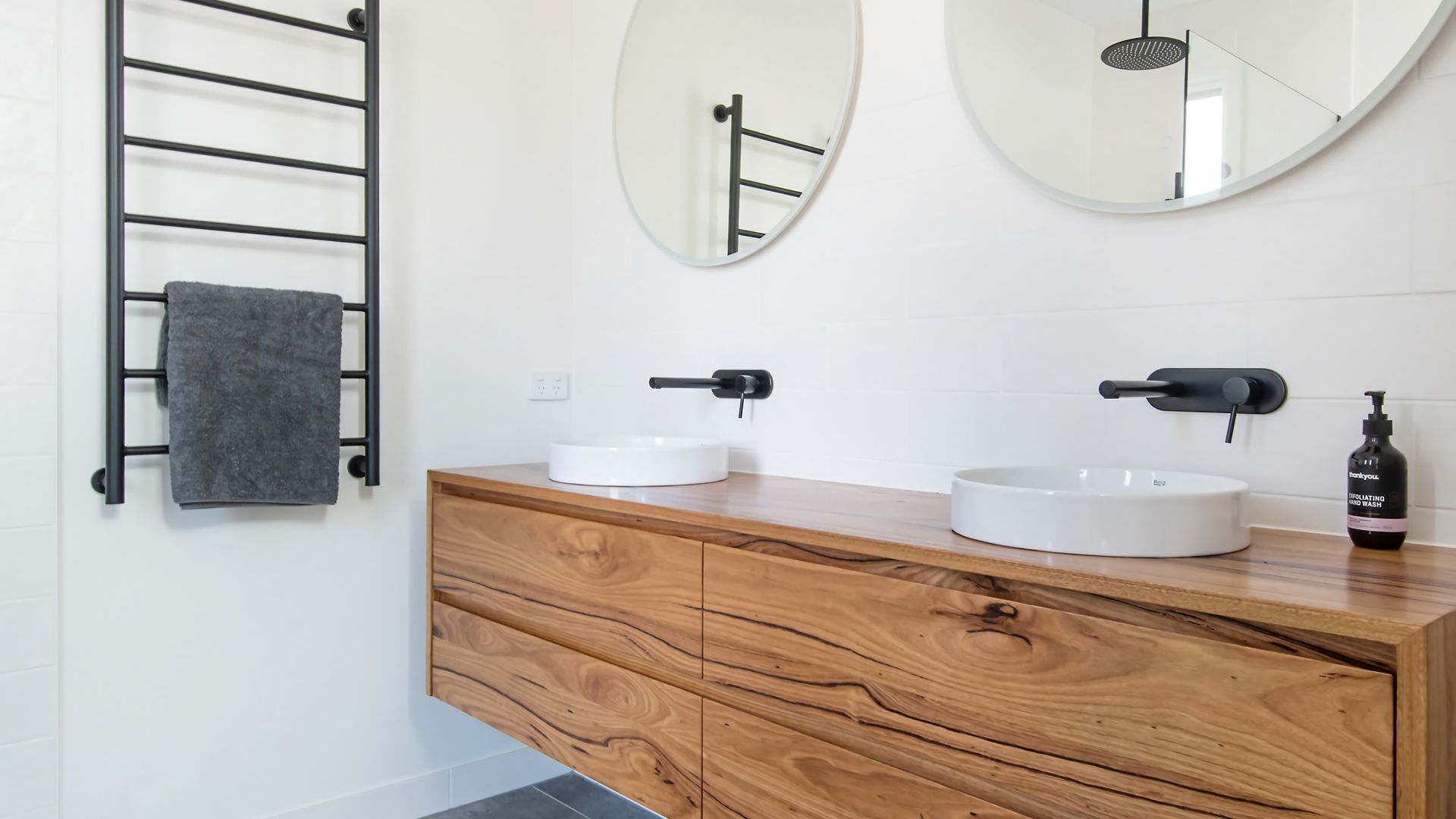 Een badkamer met een houten wastafelmeubel, twee ronde spiegels, een handdoekrek en een fles handzeep.