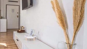 Una sala moderna con un sofá blanco, un televisor de pantalla plana montado en la pared, una pequeña mesa redonda con un jarrón de plumas de pampa secas y suelo de madera.
