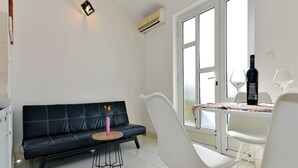 Un espacio habitable moderno con un sofá de cuero negro, una silla blanca, una mesita redonda con una botella y vasos, y un amplio ventanal con vistas.