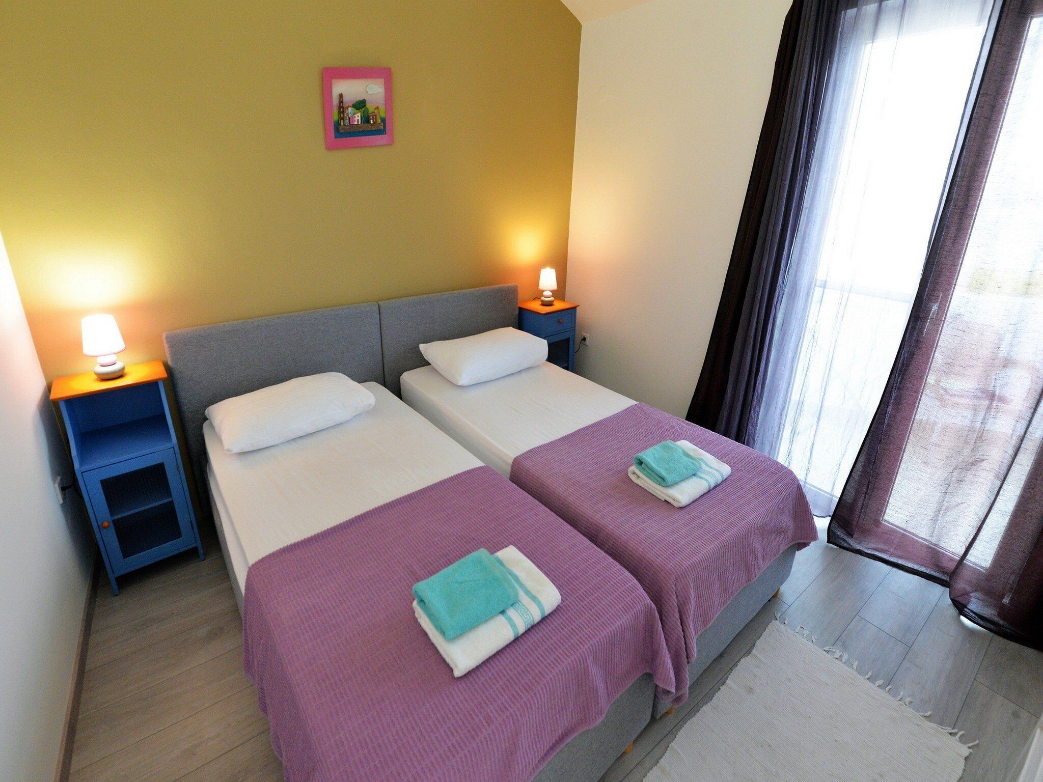 A room with two beds, nightstands, a lamp, a picture on the wall, and a window with curtains.