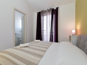 A hotel room with a bed, a nightstand with a lamp, a window with curtains, and a bathroom visible through an open door.