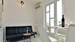 Una sala de estar moderna con un sofá de cuero negro, una silla blanca, una mesita redonda y una mesa de comedor con dos copas de vino y una botella.
