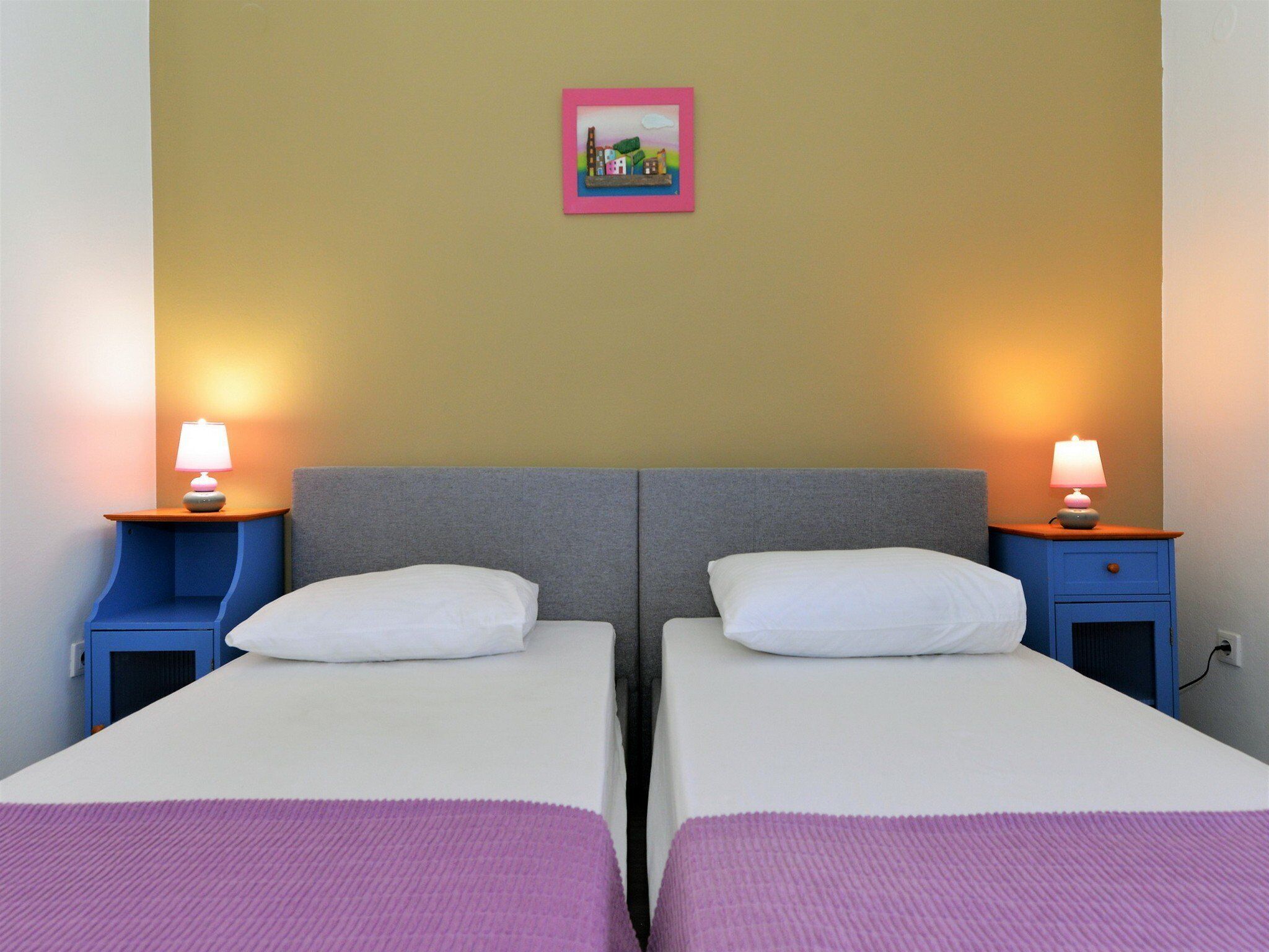 Ein Doppelzimmer mit zwei Betten, jeweils mit einem Kopfkissen und einer violetten Decke. Es gibt zwei Nachttische mit Lampen und ein kleines gerahmtes Bild an der Wand.