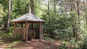Ein hölzerner Pavillon mit einem Reetdach, umgeben von dichtem Wald.
