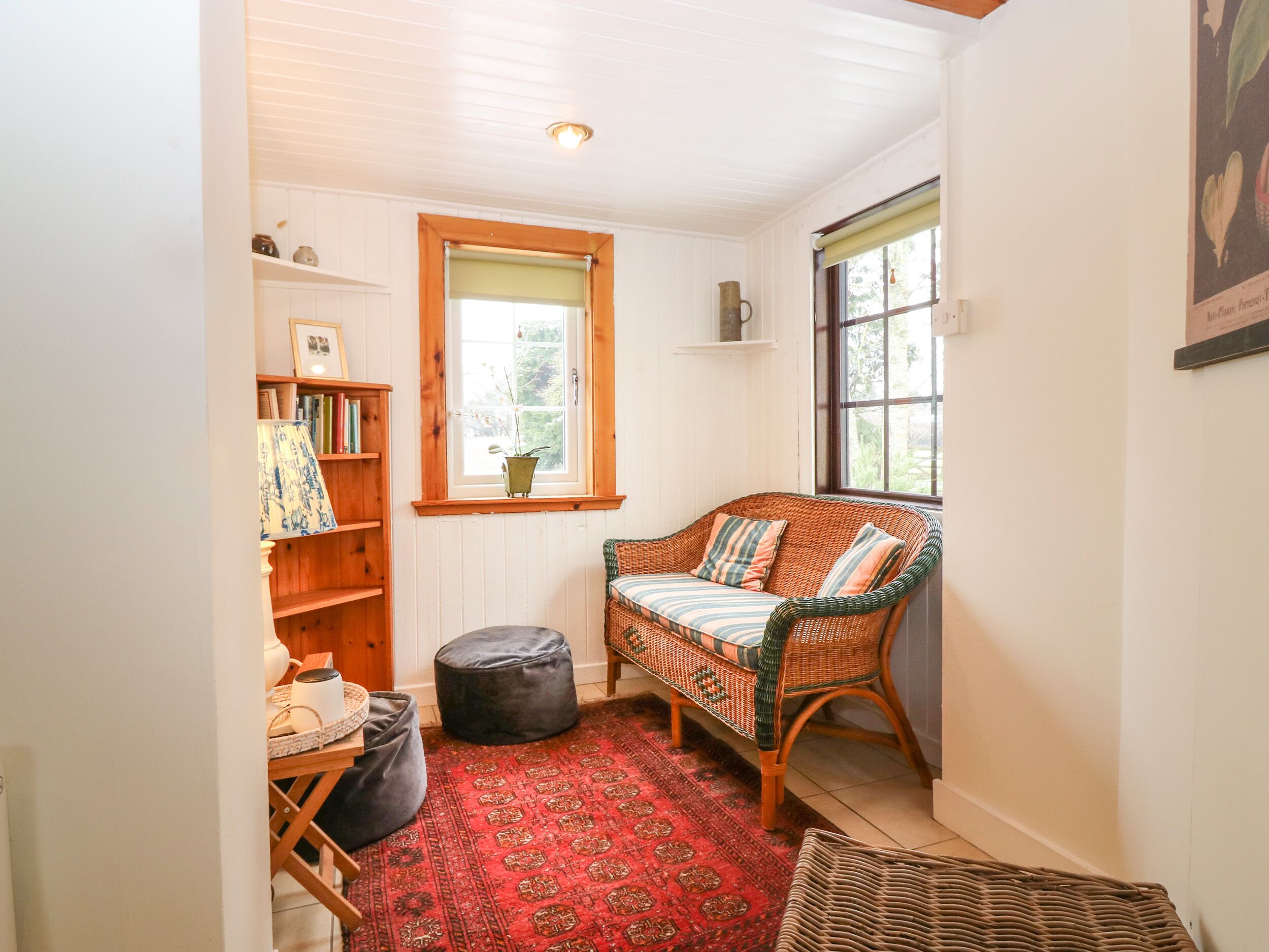 A cozy room with a wicker sofa, a small rug, a bookshelf, and a window with a view of greenery.