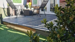 Un patio con mobiliario exterior y una pequeña mesa, rodeado por un muro de ladrillo y una puerta de cristal que da a un jardín.