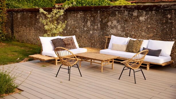 Un espace extérieur aménagé avec une terrasse en bois, deux canapés blancs avec des coussins et deux fauteuils en osier.