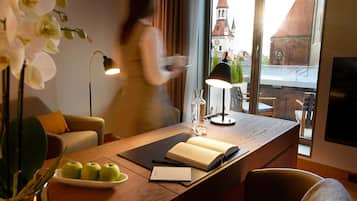 Quarto de hotel com escrivaninha de madeira, um laptop, um livro e um prato de maçãs. Há uma janela com vista para uma torre de igreja.