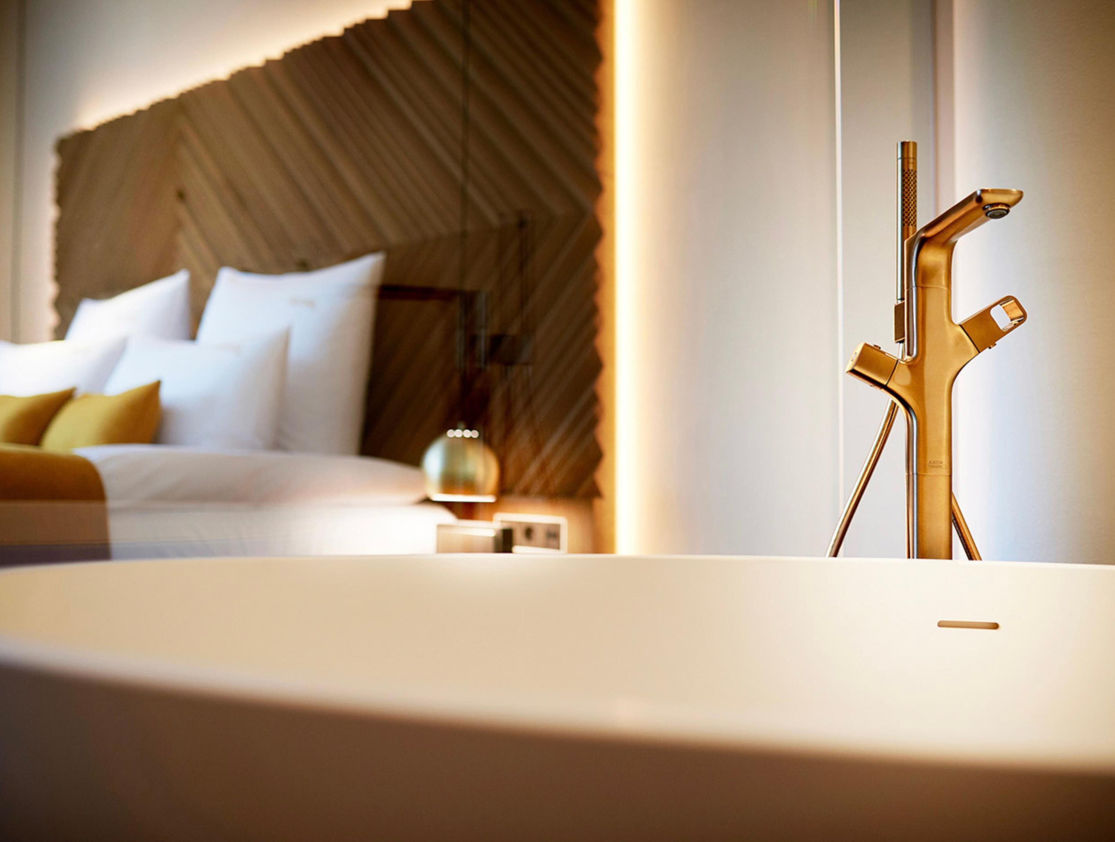 Une chambre d’hôtel moderne équipée d’un lit, d’une table de chevet, d’une lampe et d’un robinet au design unique, de couleur dorée.