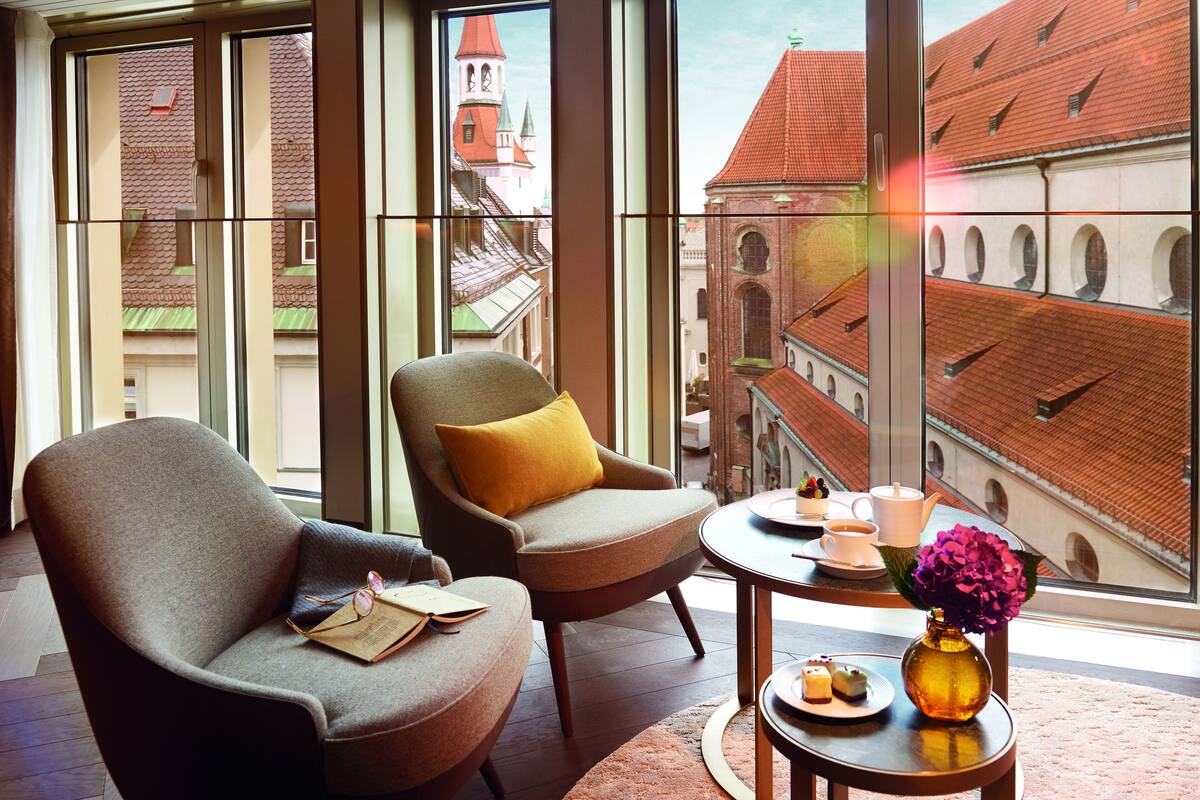 Et moderne hotelværelse med store vinduer, to lænestole, et lille rundt bord med en vase blomster og udsigt til historiske bygninger.