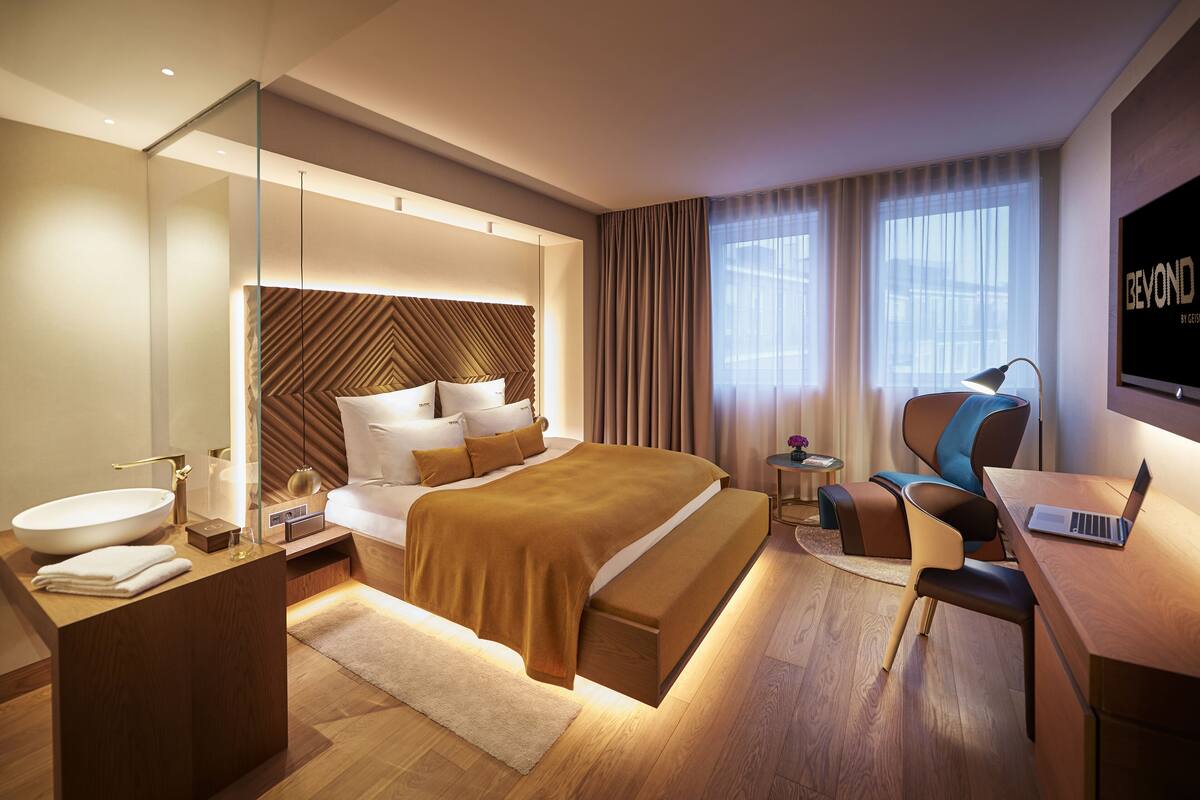 Et moderne hotelværelse med en stor seng, et skrivebord, en bærbar computer, et badeværelse med en stor vask og et fjernsyn på væggen.