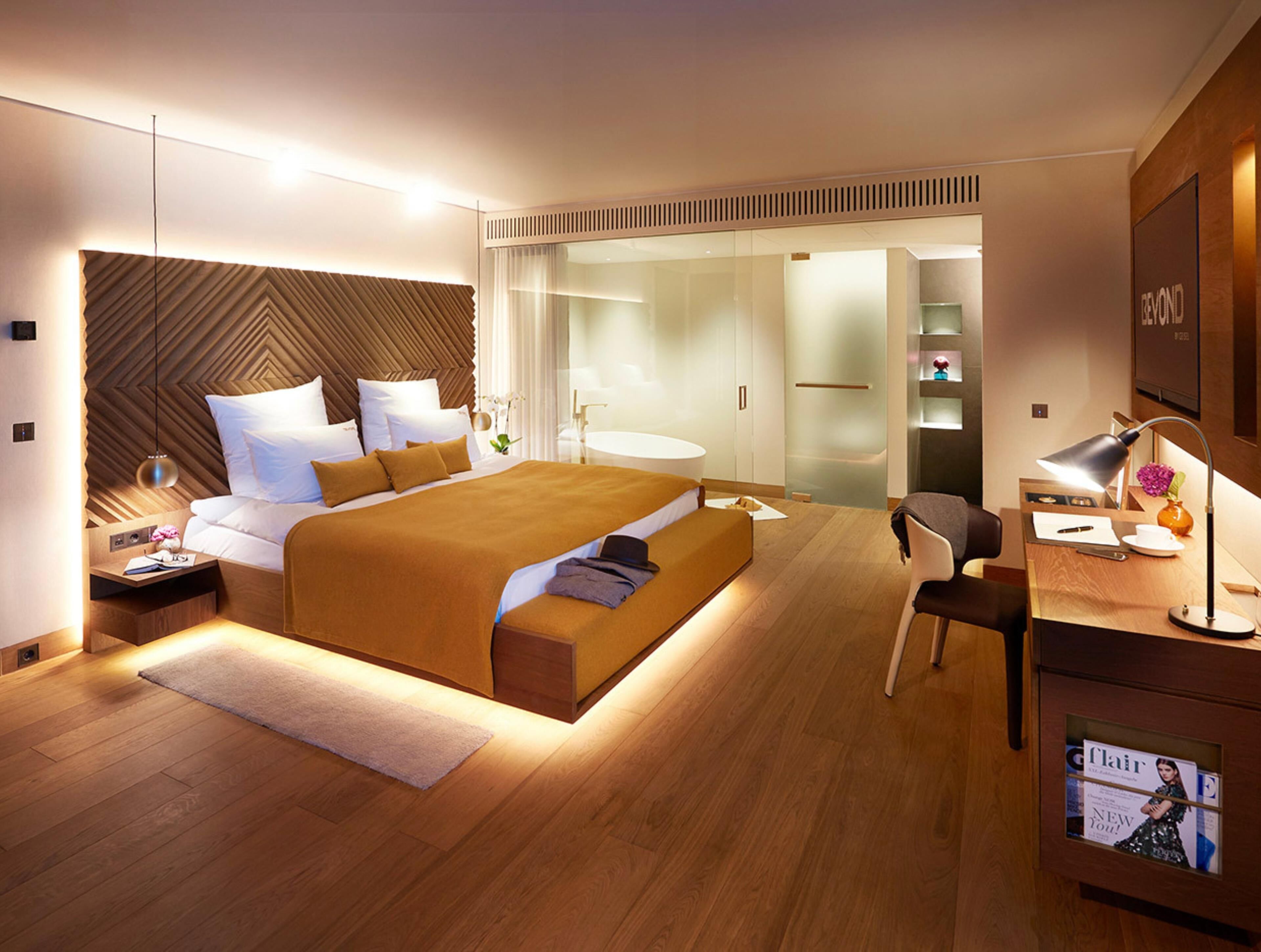 Une chambre moderne avec un grand lit, des tables de chevet, un bureau avec une chaise et une salle de bain séparée par une cloison en verre.