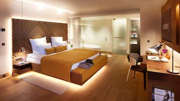 Une chambre moderne avec un grand lit, des tables de chevet, un bureau avec une chaise et une salle de bain séparée par une cloison en verre.