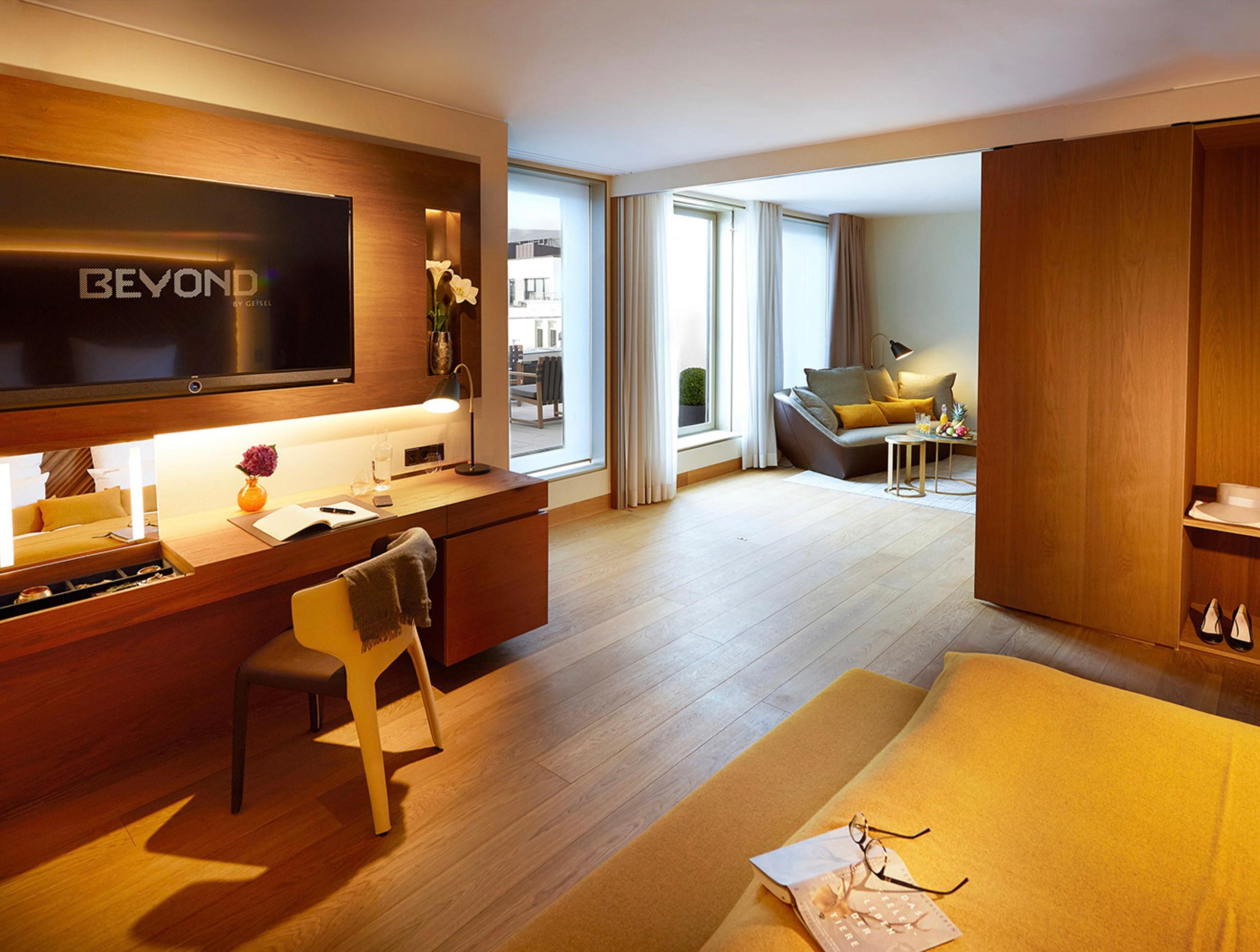Une chambre d’hôtel moderne équipée d’une télévision à écran plat, d’un bureau avec une lampe, d’un canapé et d’une grande fenêtre donnant sur l’extérieur.