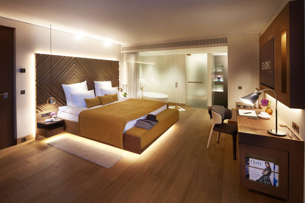 Et moderne hotelværelse med en stor seng, sengeborde, et skrivebord med lampe, et fjernsyn og et badeværelse, der kan ses gennem en glasvæg.