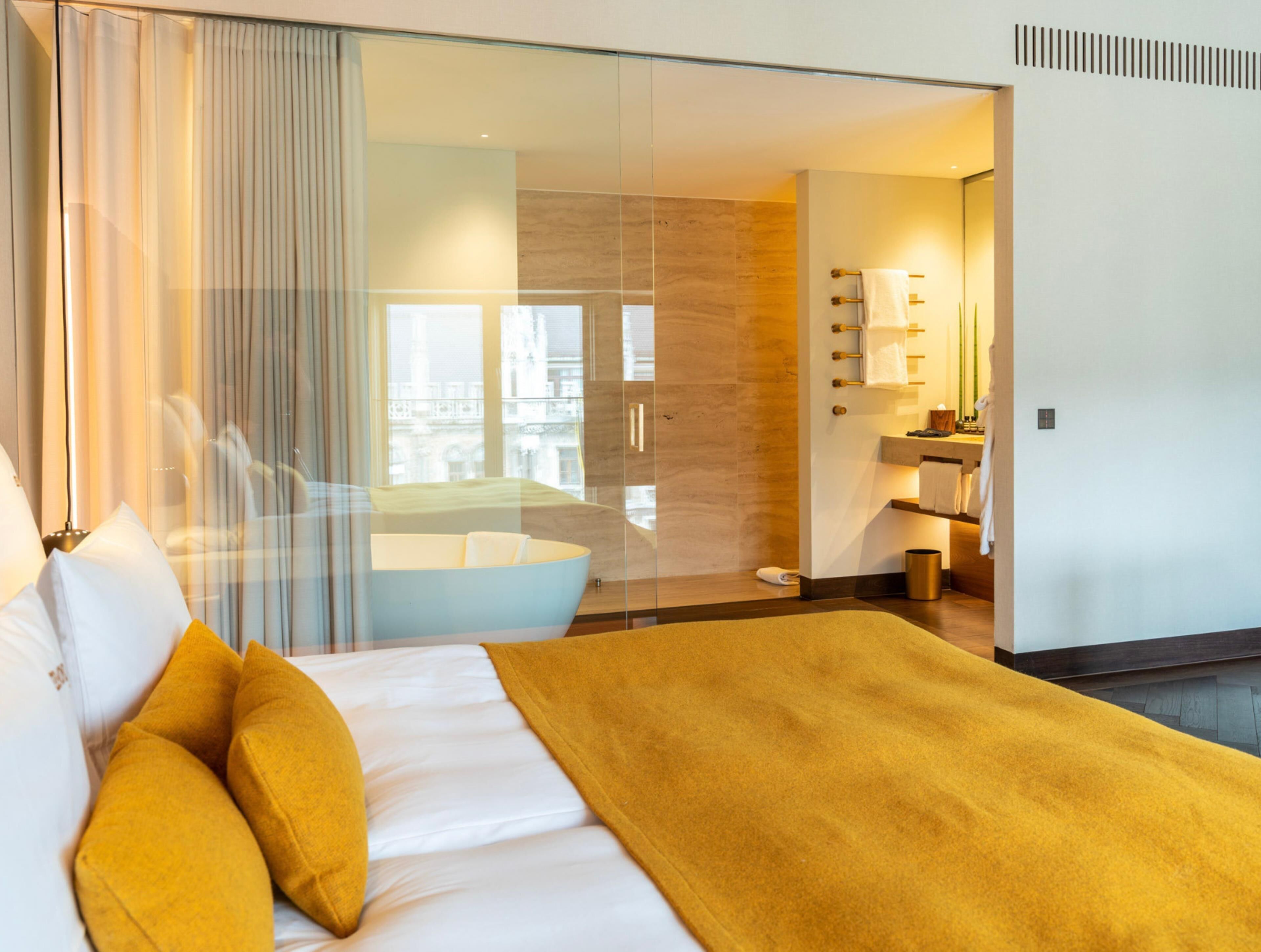 A modern hotel room with a large bed, a glass partition, a bathroom with a bathtub, and a city view.