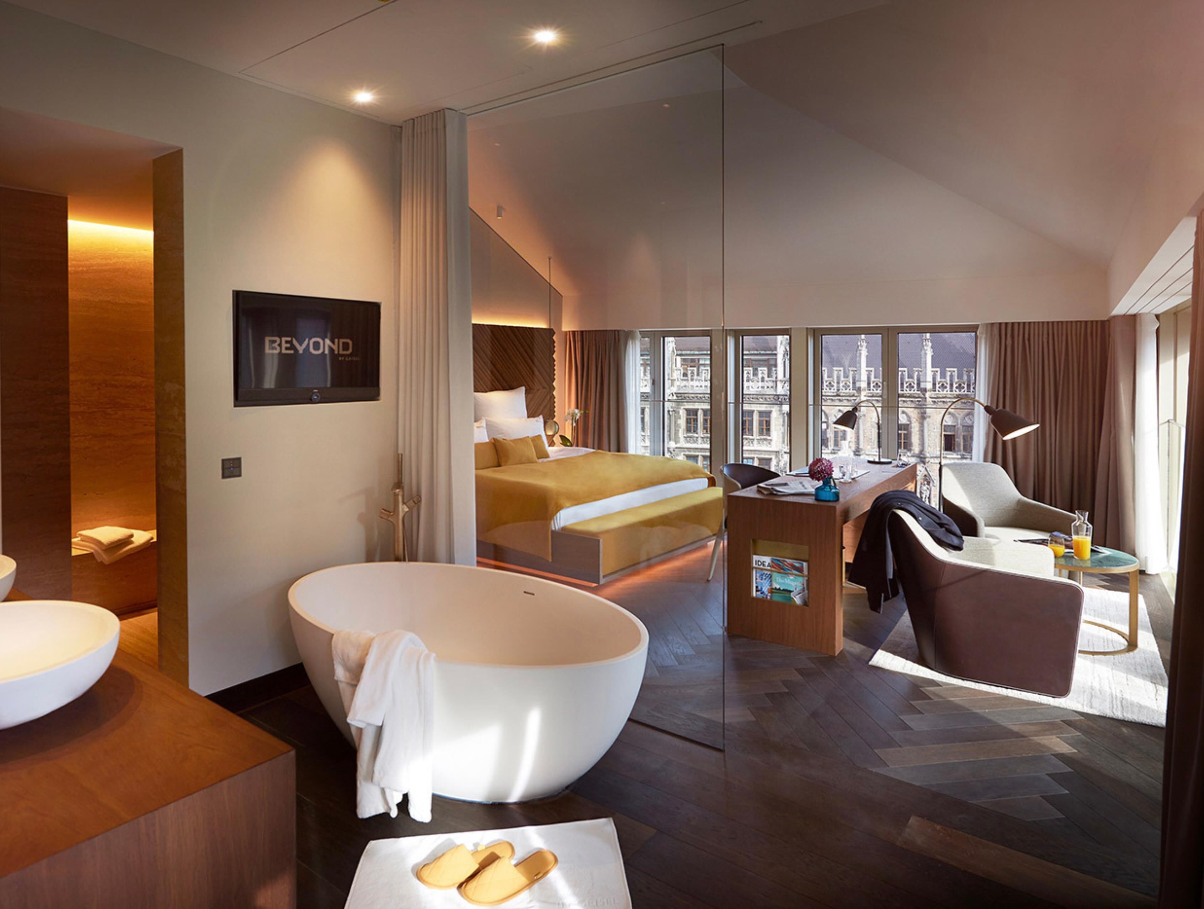 Une chambre d’hôtel moderne dotée d’une grande baignoire, d’un lit avec vue, d’un bureau avec une lampe et d’une vue sur la ville à travers les fenêtres.