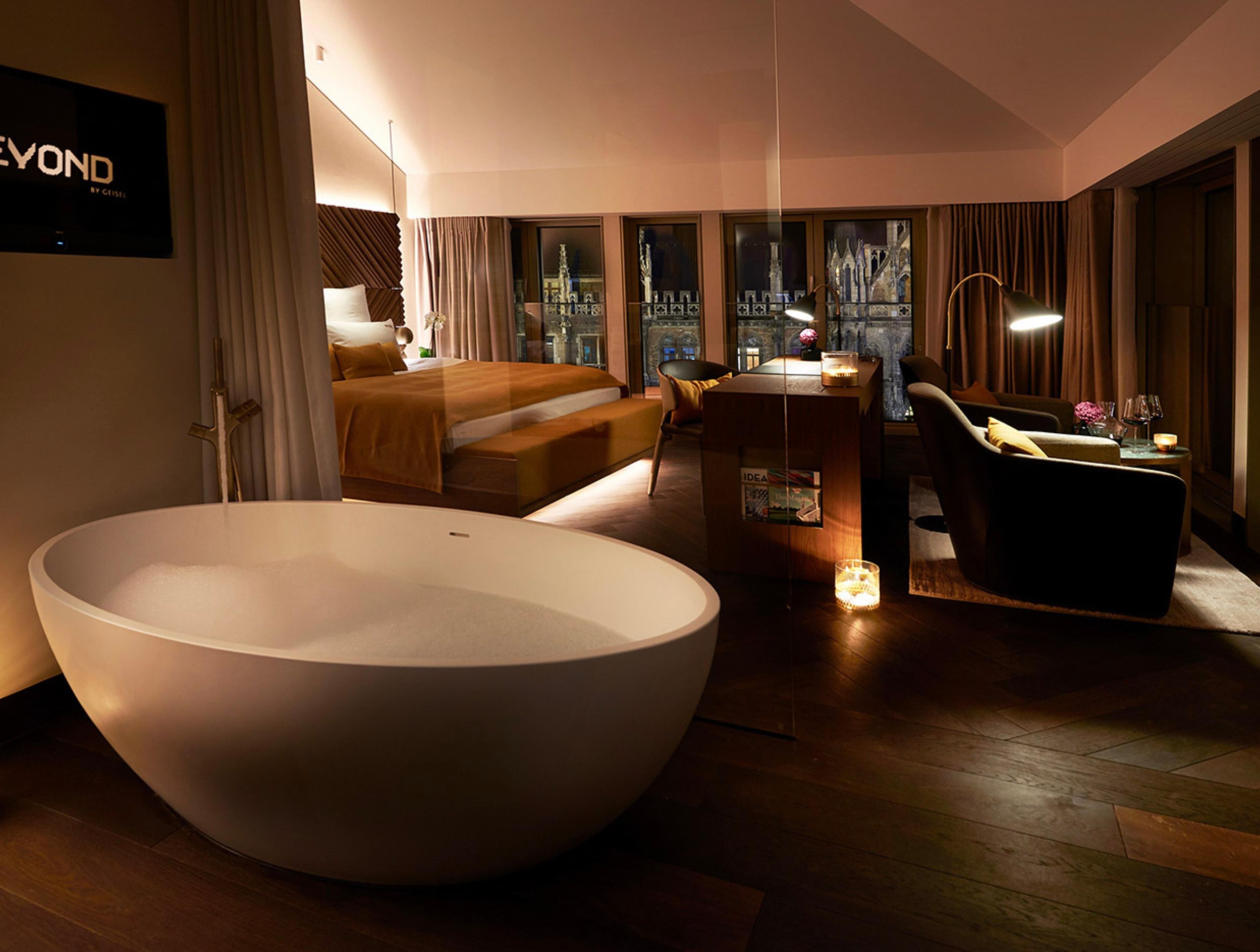 A modern hotel room with a large bathtub, a bed, a desk, and a view of the cityscape through large windows.